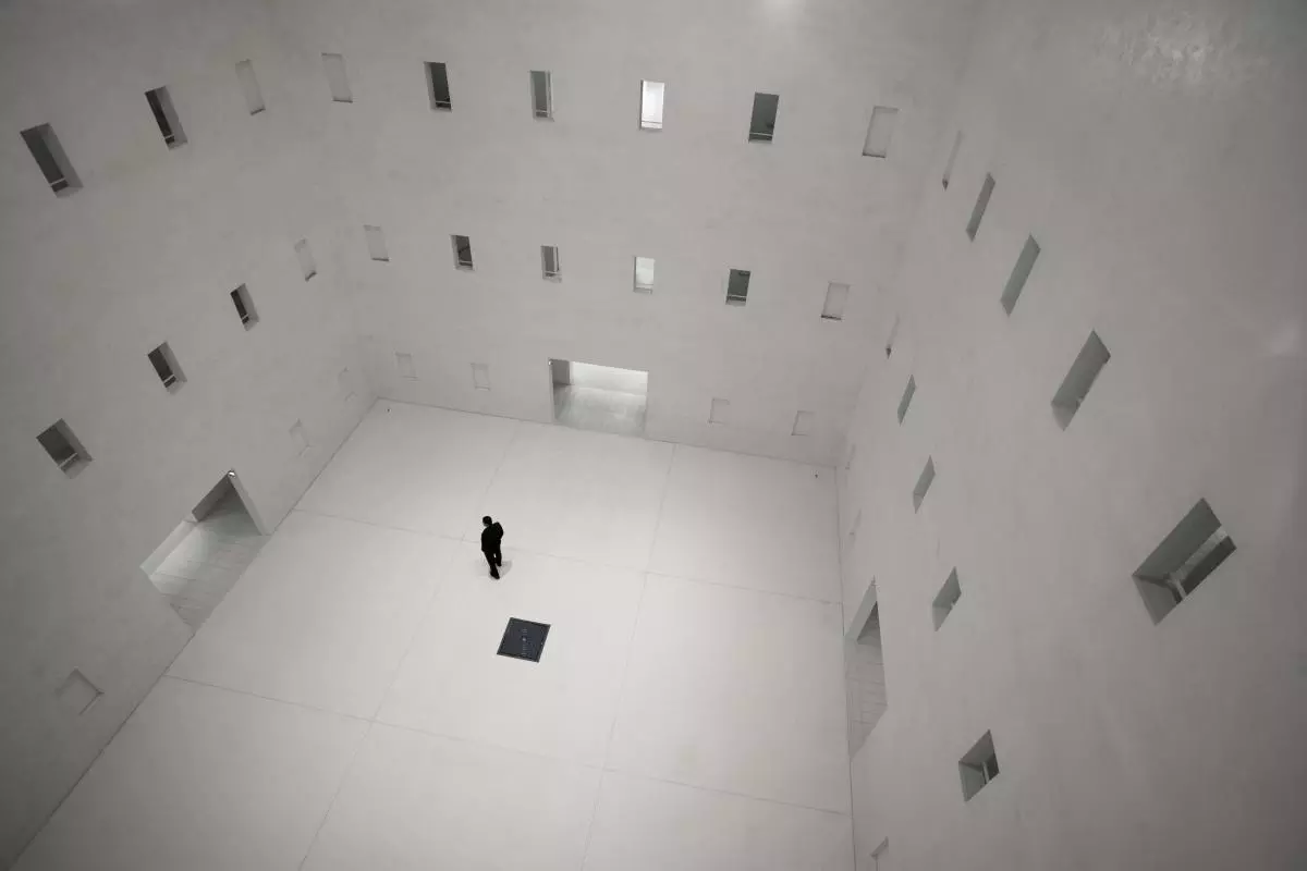 A figure stands in the middle of a room with tall walls containing multiple doors and windows on every side 